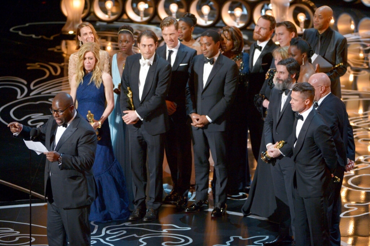 Image displaying Director Steve McQueen and the cast and crew of 12 Years a Slave stand on stage at the Oscars, dressed in formal attire, as they accept the Best Picture award.