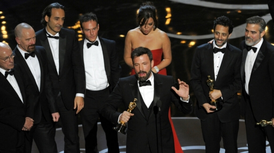 Ben Affleck stands at the podium holding an Oscar while delivering a speech, surrounded by the cast and crew of Argo on stage at the 2013 Academy Awards. The group, dressed in formal attire, celebrates their Best Picture win at the Dolby Theatre.