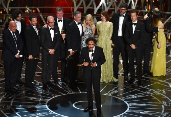 Director Alejandro G. Iñárritu stands at the microphone holding an Oscar, delivering a speech as the cast and crew of Birdman or (The Unexpected Virtue of Ignorance) celebrate on stage at the 2015 Academy Awards. The group, dressed in formal attire, smiles and applauds behind him on the ornate stage of the Dolby Theatre in Los Angeles.